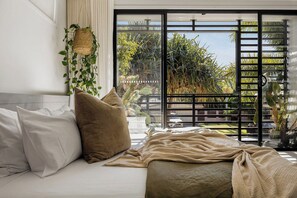 Een slaapkamer met een bed, kussens, een hangplant en uitzicht door een raam met een zwart metalen hek.