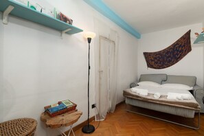 Een slaapkamer met een bed, een staande lamp, een klein tafeltje met boeken, een geweven stoel en een blauwe plank met spullen.