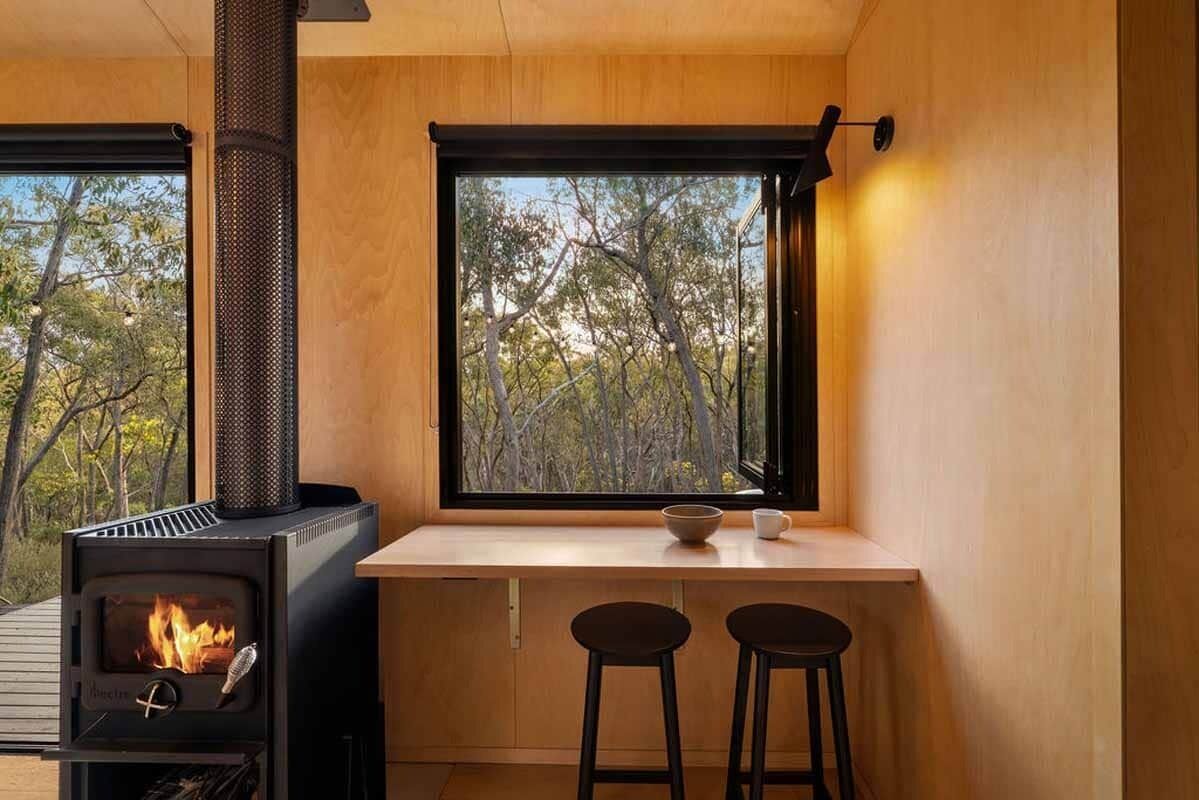 A modern kitchen with a wood-burning stove, a window with a view of trees, and two stools.