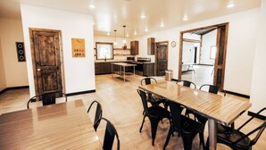 A modern dining area with a wooden table and chairs, a kitchen in the background, and a large mirror on the wall.