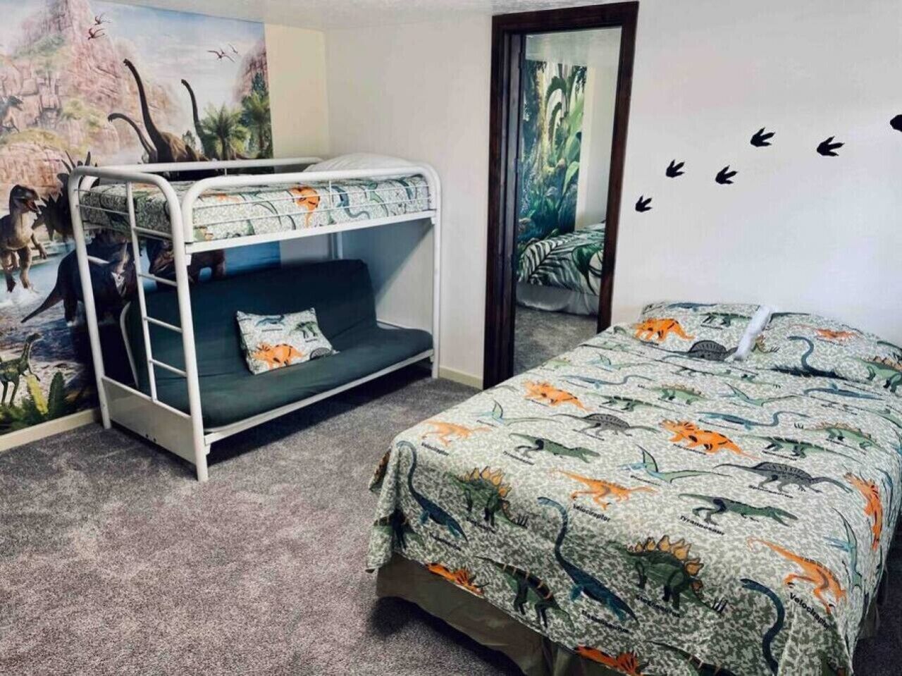 Ein Zimmer mit Etagenbett, einem Bett mit Bettwäsche im Dinosaurer-Design, einem Wandbild mit Dinosauriern und einem Spiegel mit einer Naturdarstellung.