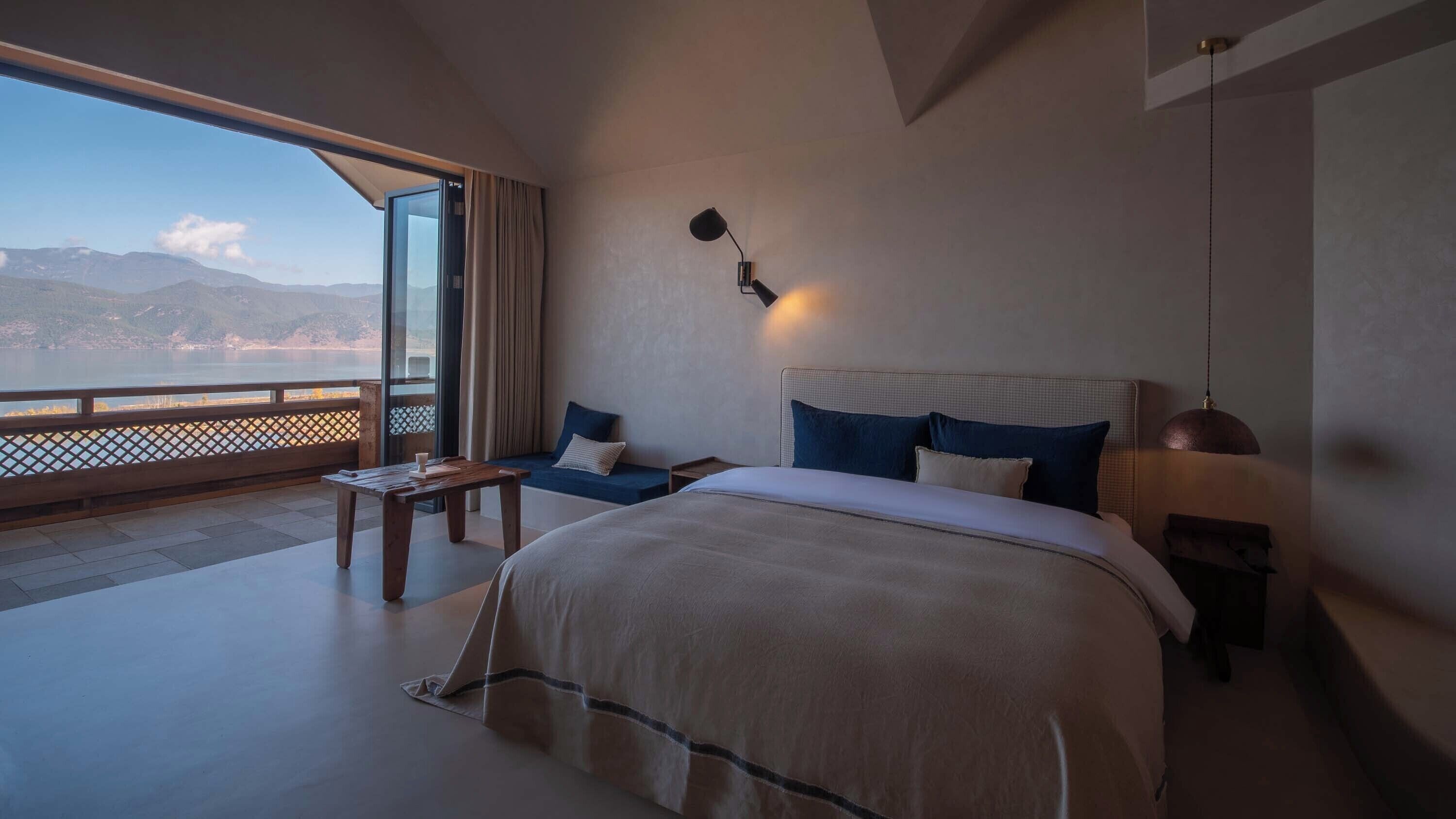 Une chambre avec un grand lit, une table de chevet, une lampe et une vue sur l’extérieur.