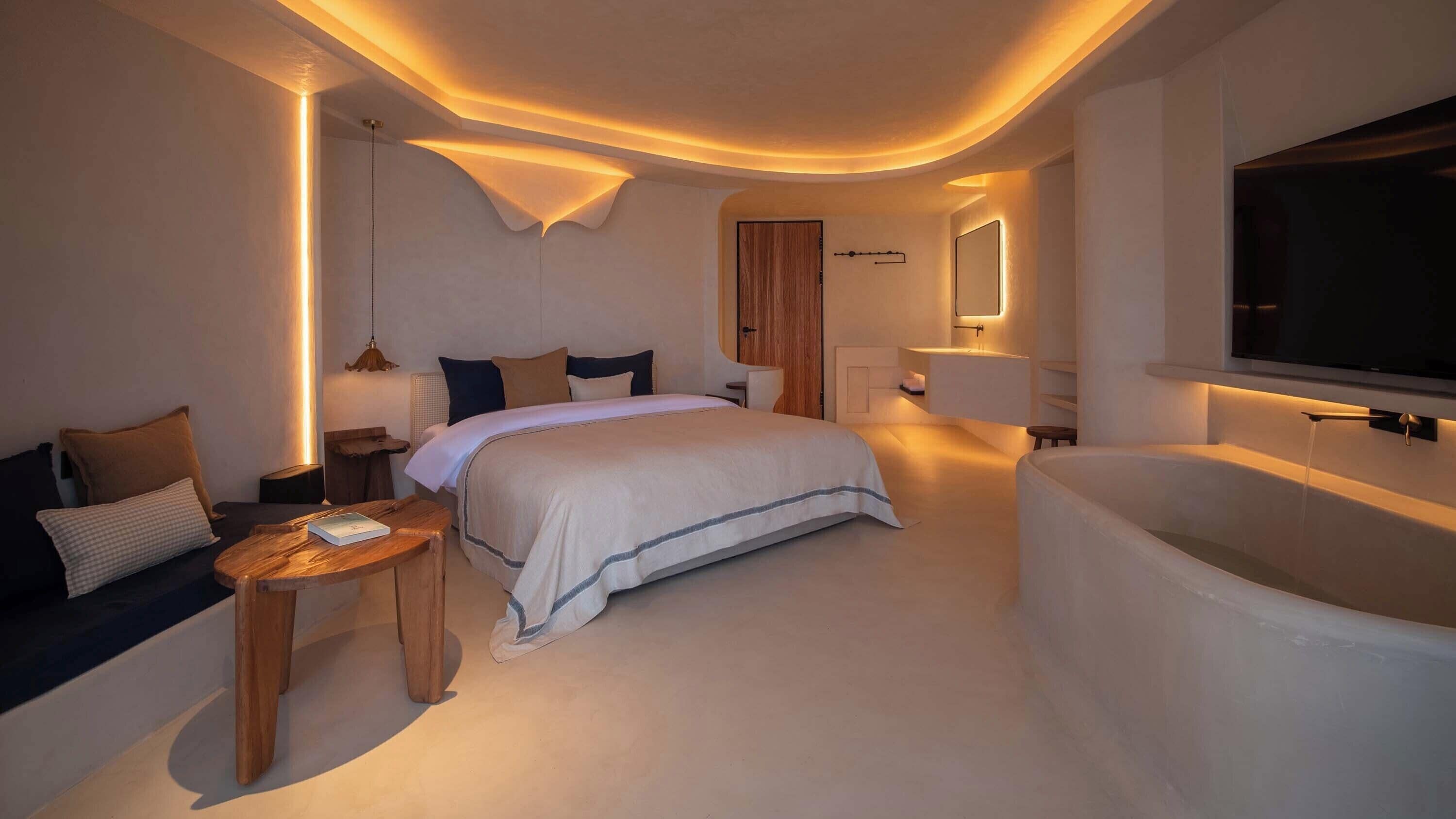 Ein modernes Hotelzimmer mit einem großen Bett, einer Sitzecke mit kleinem Tisch, einer integrierten Badewanne und einem an der Wand befestigten Fernseher.