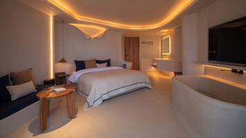 Ein modernes Hotelzimmer mit einem großen Bett, einer Sitzecke mit kleinem Tisch, einer integrierten Badewanne und einem an der Wand befestigten Fernseher.