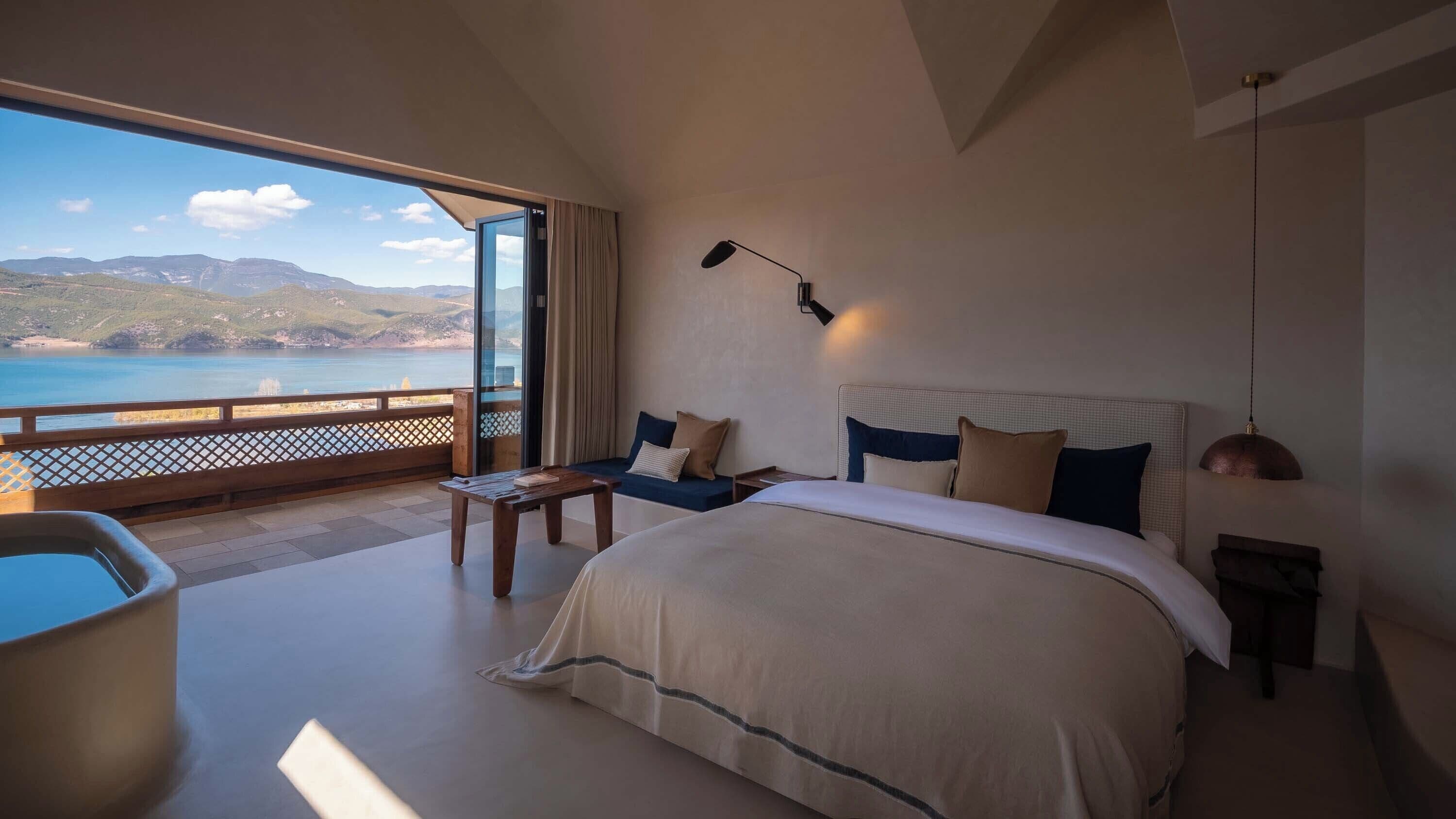 A bedroom with a large bed, a bedside table, a nightstand, a lamp, and a view of the sea.