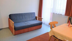 Une petite pièce avec un canapé, une table recouverte d’une nappe et une fenêtre avec des rideaux.