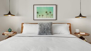 Uma cama bem arrumada com um travesseiro branco, uma moldura com uma imagem de palmeiras e duas luminárias pendentes.