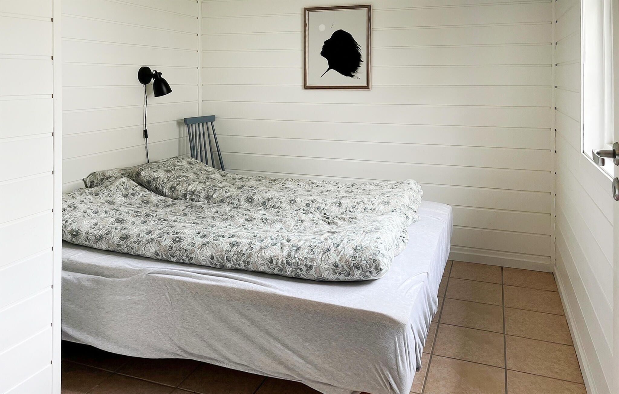Un lit simple avec une couvre-lit à motifs floraux, une lampe fixée au mur et une œuvre d’art représentant une silhouette encadrée, le tout sur un mur blanc à panneaux.