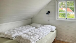 A small, single-bed room with white bedding, a sloped ceiling, a window with a view of greenery and buildings, and a black lamp. - Cozy home in Lindesnes with WiFi (Lindesnes)