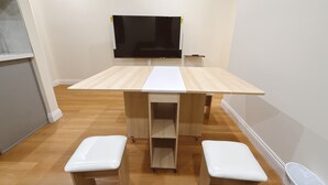 Ein Zimmer mit einem Holztisch, zwei weißen Stühlen, einem weißen Schrank und einem flachbildfernseher an der Wand.