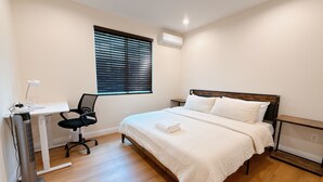 Ein Schlafzimmer mit einem Bett, einem Schreibtisch mit Stuhl, einem Standventilator und einem Fenster mit Jalousien.