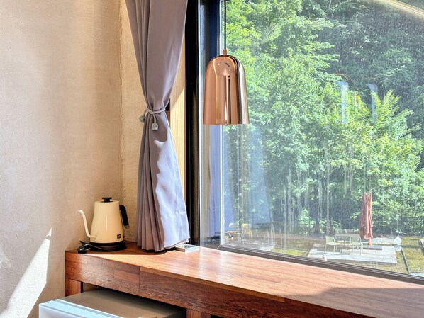 A window with a curtain, a kettle, and a view of a forest.