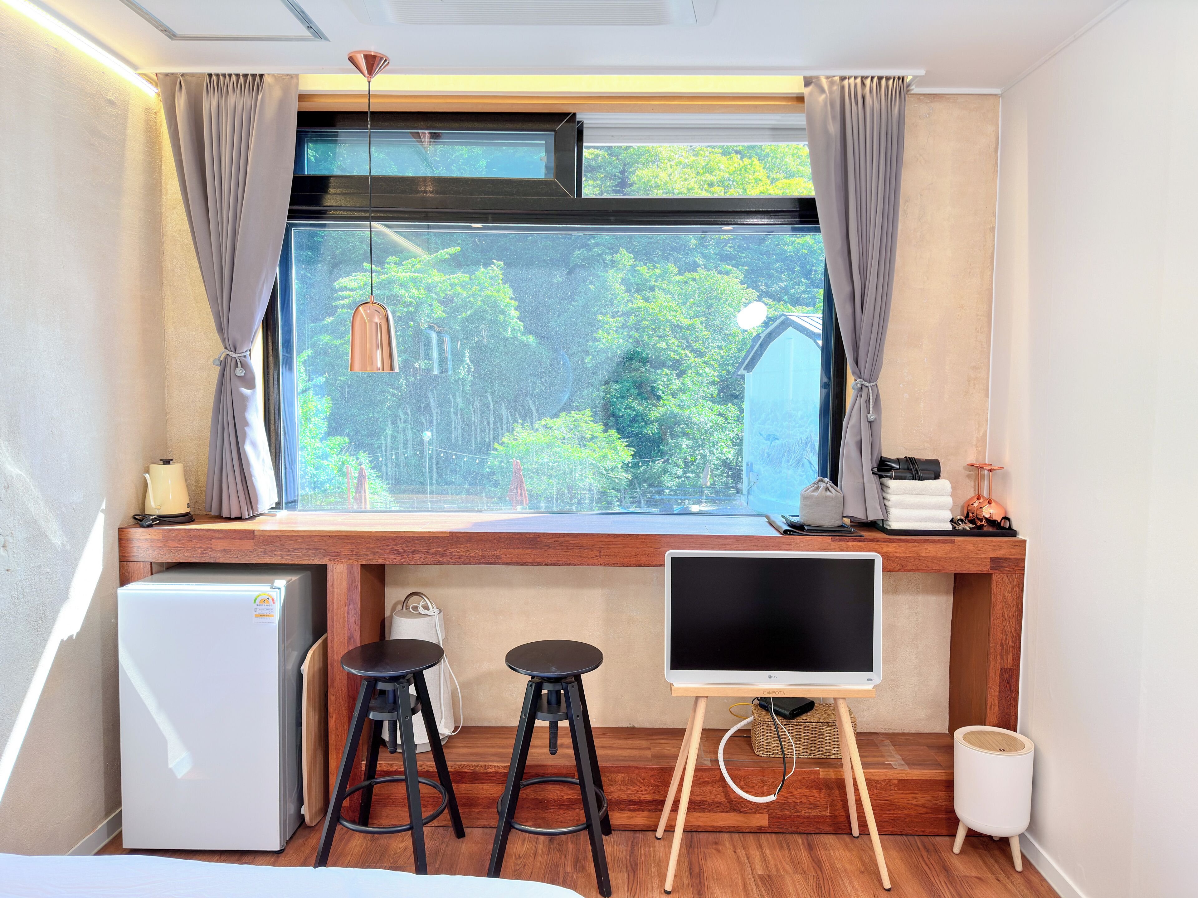 A room with a window, a small fridge, two stools, a TV on a stand, and a waste bin.