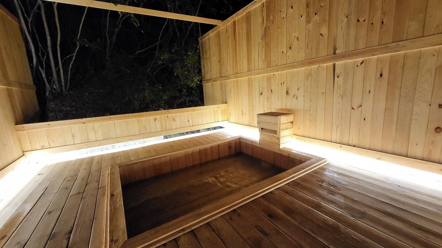 A wooden sauna with a rectangular hot tub, wooden walls, and a window overlooking a forest.