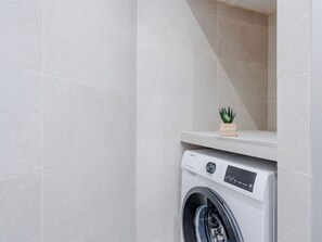 A modern laundry room with a front-loading washing machine, a small wooden shelf with a plant, and light-colored tiled walls. - Apartment Cofisa by Interhome (Jávea)