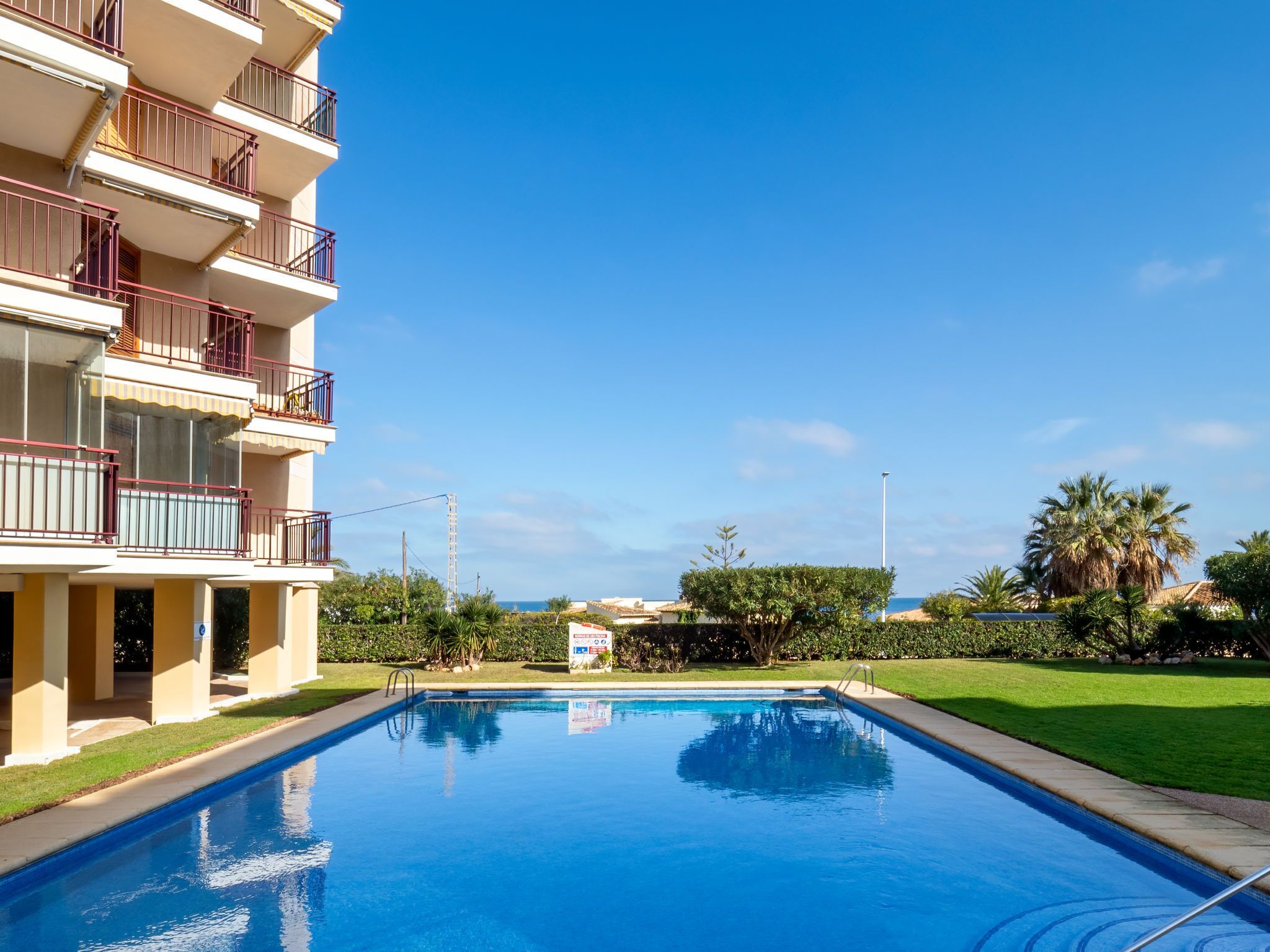 Una piscina con un cielo azul despejado, un edificio de varios pisos con balcones y un césped bien cuidado.