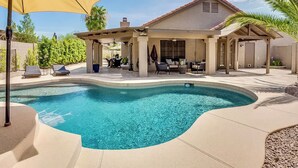 Uma piscina com borda curva, um espaço coberto com área de estar e uma casa com telhado inclinado.