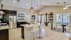 Uma cozinha moderna com eletrodomésticos de aço inoxidável, uma ilha central com banquetas, iluminação pendente e uma área de jantar com mesa e cadeiras de vidro.