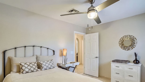 Quarto com cama, mesa de cabeceira, cômoda, ventilador de teto e porta.