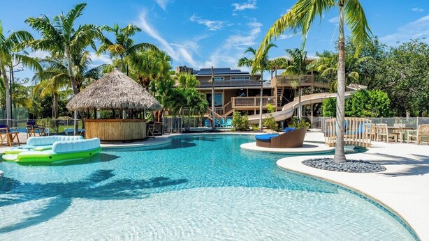 A swimming pool with lounge chairs, a thatched-roof hut, and a building with a balcony. - Villa Zephyra, Grand 8BR Villa for Families & Groups (Jensen Beach)