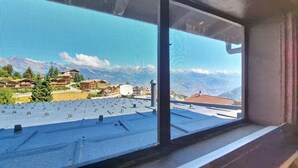 Une fenêtre donnant sur un paysage montagneux, un bâtiment avec un toit rouge et un ciel dégagé.