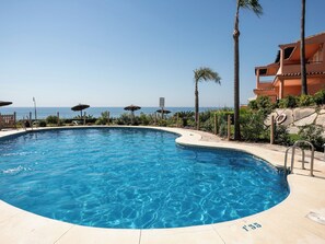 A clear blue pool with a curved edge, surrounded by palm trees and a view of the sea. - Urbanización Bermuda Beach by Interhome (Estepona)