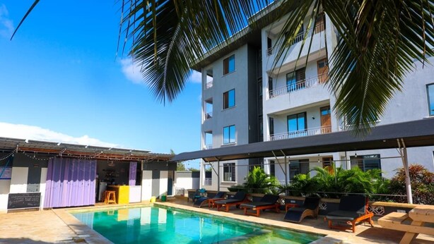Una piscina con sillones de descanso y zona de bar, rodeada por un edificio de varios pisos con balcones.