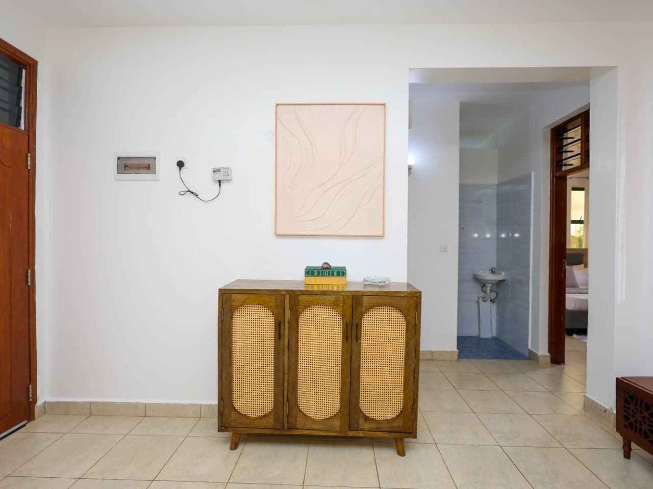 A room with a wooden cabinet, a framed artwork, a wall socket, and a bathroom visible through an open door.