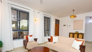 Una sala de estar con un sofá, dos sillones, una mesa de centro y una mesa de comedor. Hay cortinas, un aparador de madera y una lámpara fijada a la pared.