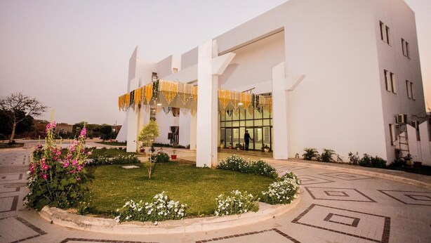 A modern building with a white facade, a central entrance with glass doors, and a landscaped garden area with flowers and a small tree. - MEERA MADHAV RESORT (Mathura)