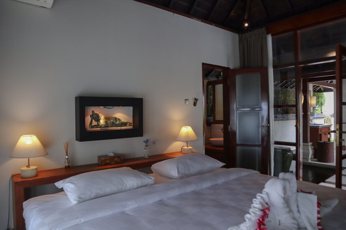 Ein Hotelzimmer mit einem Bett, zwei Nachttischlampen, einem gerahmten Bild an der Wand und einer Tür zu einem Außenbereich.