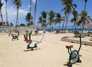 Fitnessgeräte im Freien an einem sandigen Strand mit Palmen und blauem Himmel.
