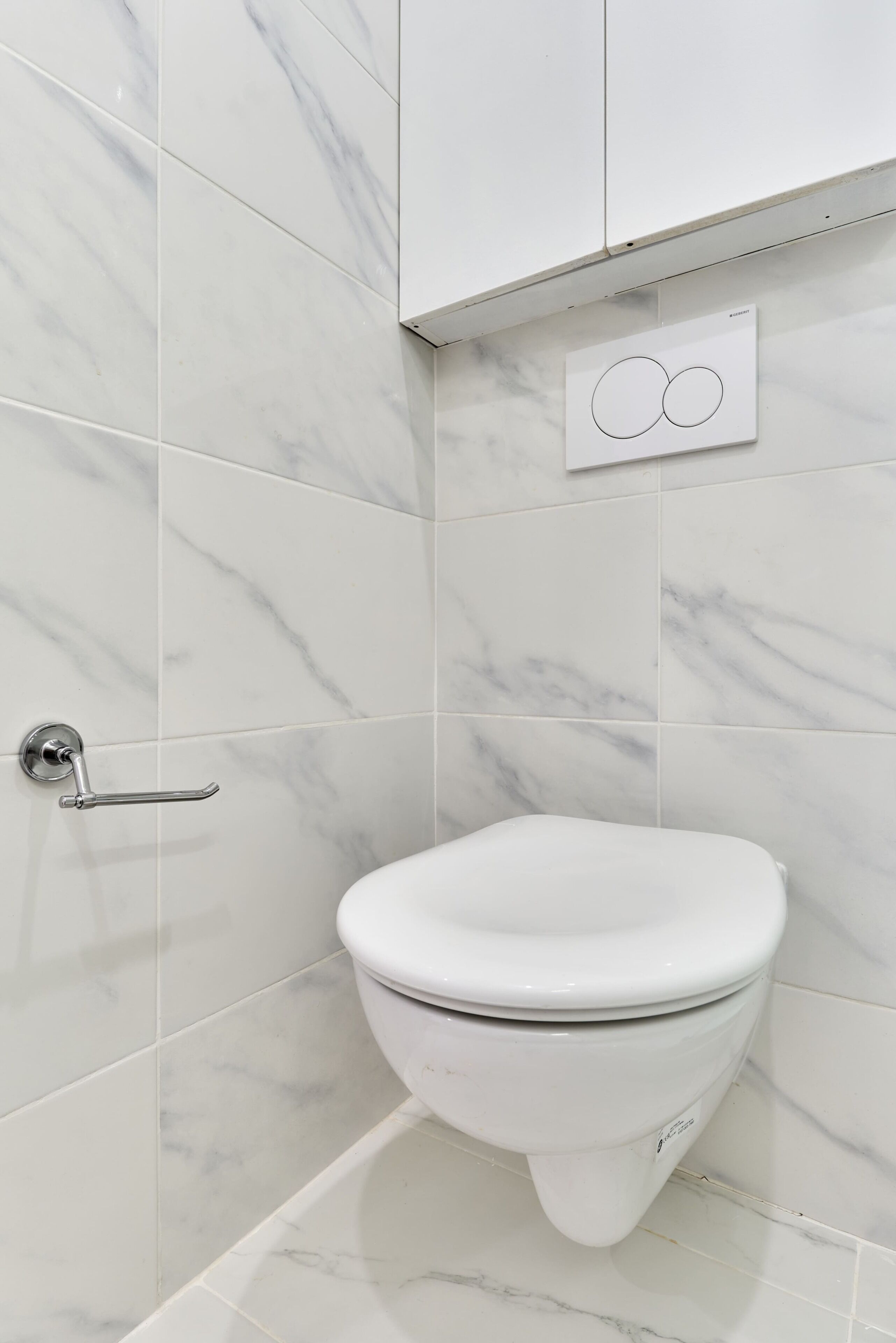 A bathroom with a toilet, white marble tiles, a wall-mounted flush handle, and a flush control panel.