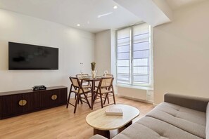 Ein modernes Wohnzimmer mit Flachbildfernseher, einem Esstisch mit Stühlen, einer Couch und einem Fenster mit Jalousien.