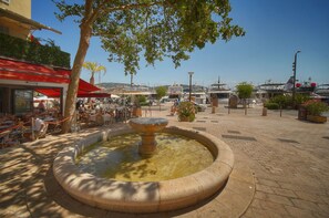 Ein öffentlicher Platz mit einem zentralen Brunnen, Sitzgelegenheiten im Freien und Booten im Hintergrund.