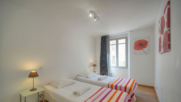 Una habitación con dos camas, una mesita de noche con lámpara, una ventana con cortinas y un cuadro de amapolas en la pared.