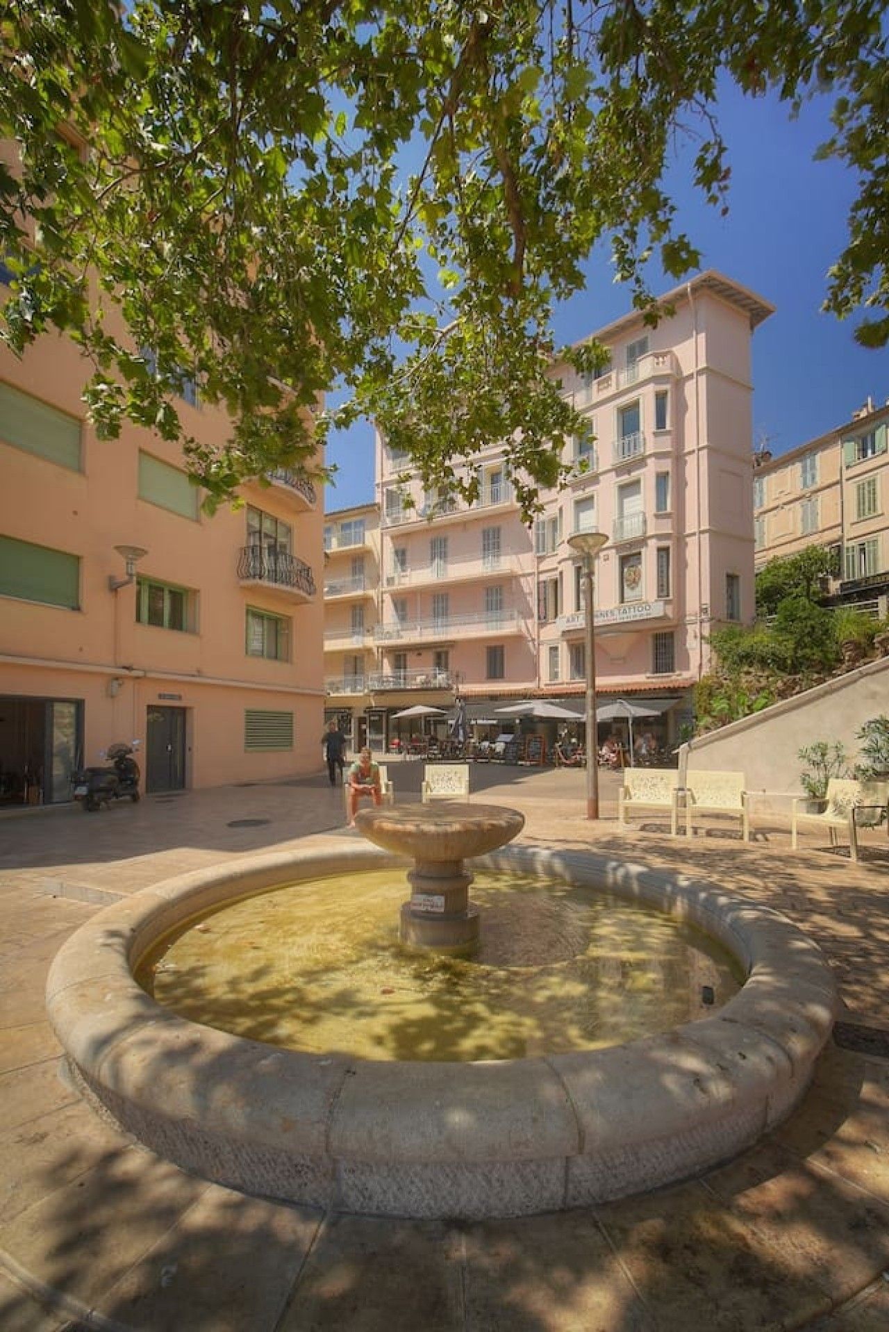 A courtyard with a central fountain, surrounded by multi-story buildings with balconies and outdoor seating areas.