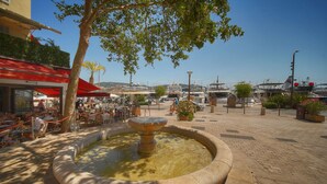 Una plaza pública con una fuente central, mesas al aire libre y barcos al fondo.
