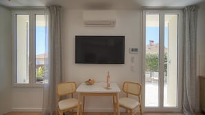 Een kamer met een flatscreen televisie, twee rieten stoelen, een klein tafeltje met een kom en een fles, en grote ramen voorzien van gordijnen.