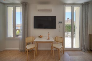 Ein Zimmer mit Flachbildfernseher, zwei Korbstühlen, einem kleinen Tisch mit Schale und Flasche sowie großen Fenstern mit Vorhängen.