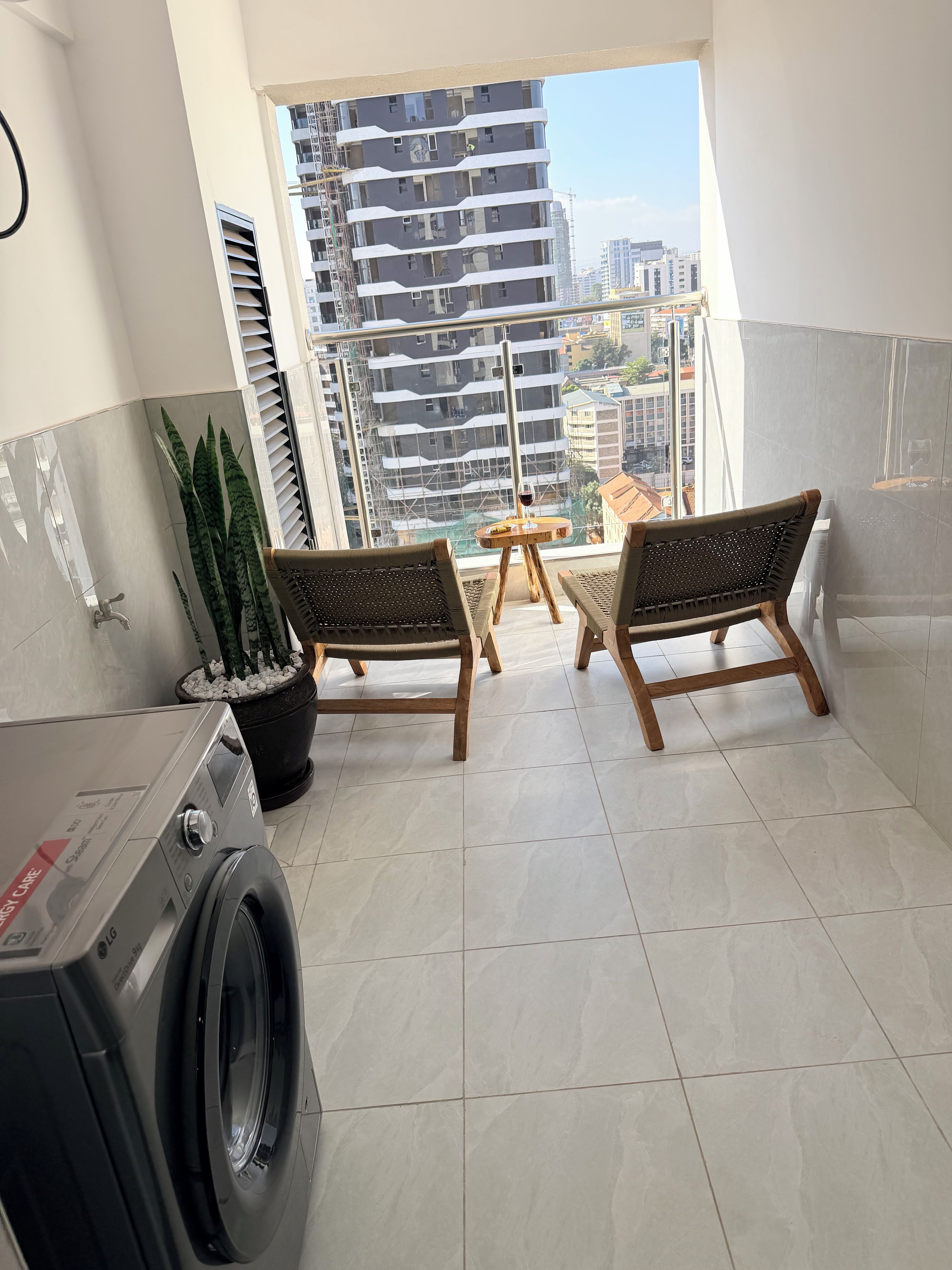 Ein Balkon mit Waschmaschine, zwei Korbstühlen, einem kleinen Tisch mit einem Weinglas und einem Topfpflanzen.