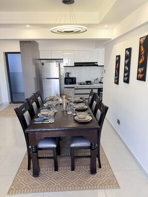 A modern dining area with a long table set for six, kitchen appliances in the background, and abstract art on the wall. - Spacious 2BR Westlands (Nairobi)
