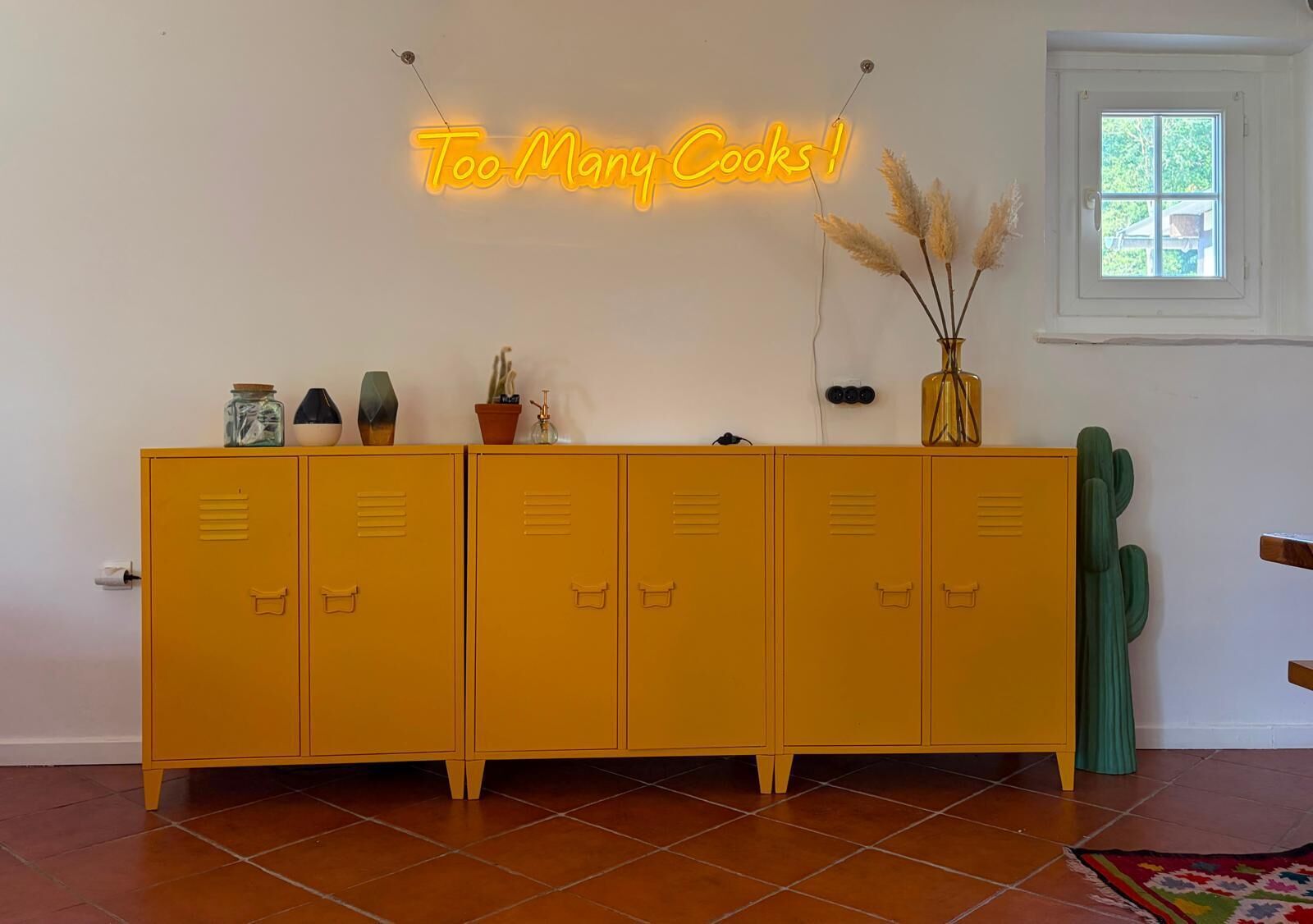 A yellow cabinet with multiple drawers, a neon sign above it, and a cactus on the right.