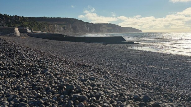 Une plage de galets, avec une digue rocheuse et des falaises en arrière-plan.