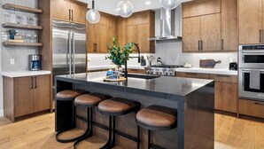Eine moderne Küche mit Holzausstattung, Edelstahlgeräten, einer zentralen Kochinsel mit Hockern, Pendelleuchten und einer Rückwand.
