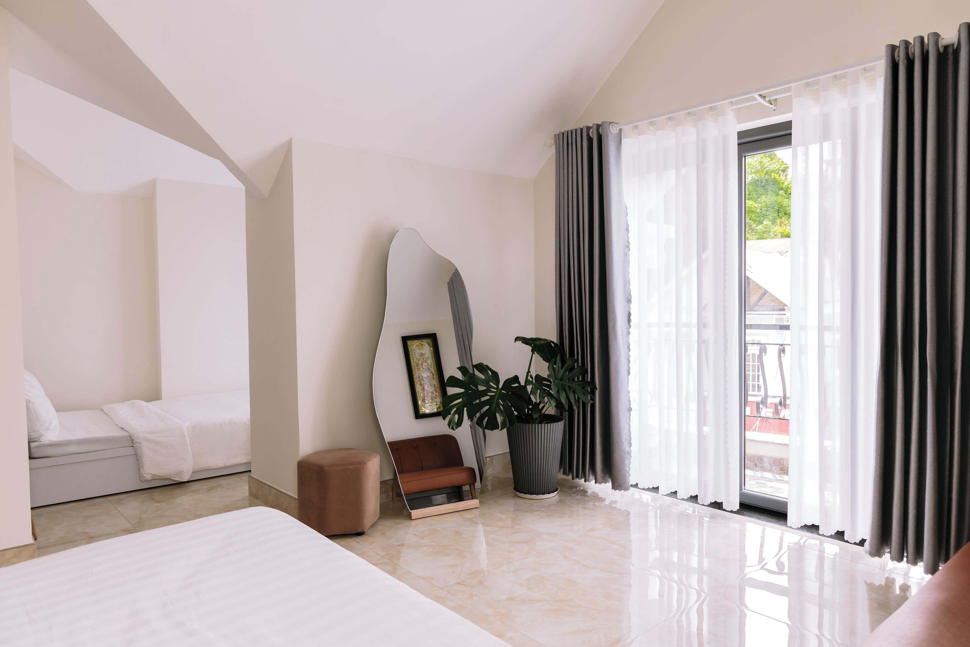Una habitación de hotel moderna con una cama, un área de descanso con un sofá y un sillón, una planta en maceta y un gran ventanal con cortinas.