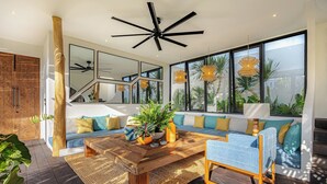 A modern living room with a wooden coffee table, blue and yellow sofa, large windows, and a ceiling fan. - Brand-New LUX 5BR Villa with Grand Pool & Garden (Kerobokan)