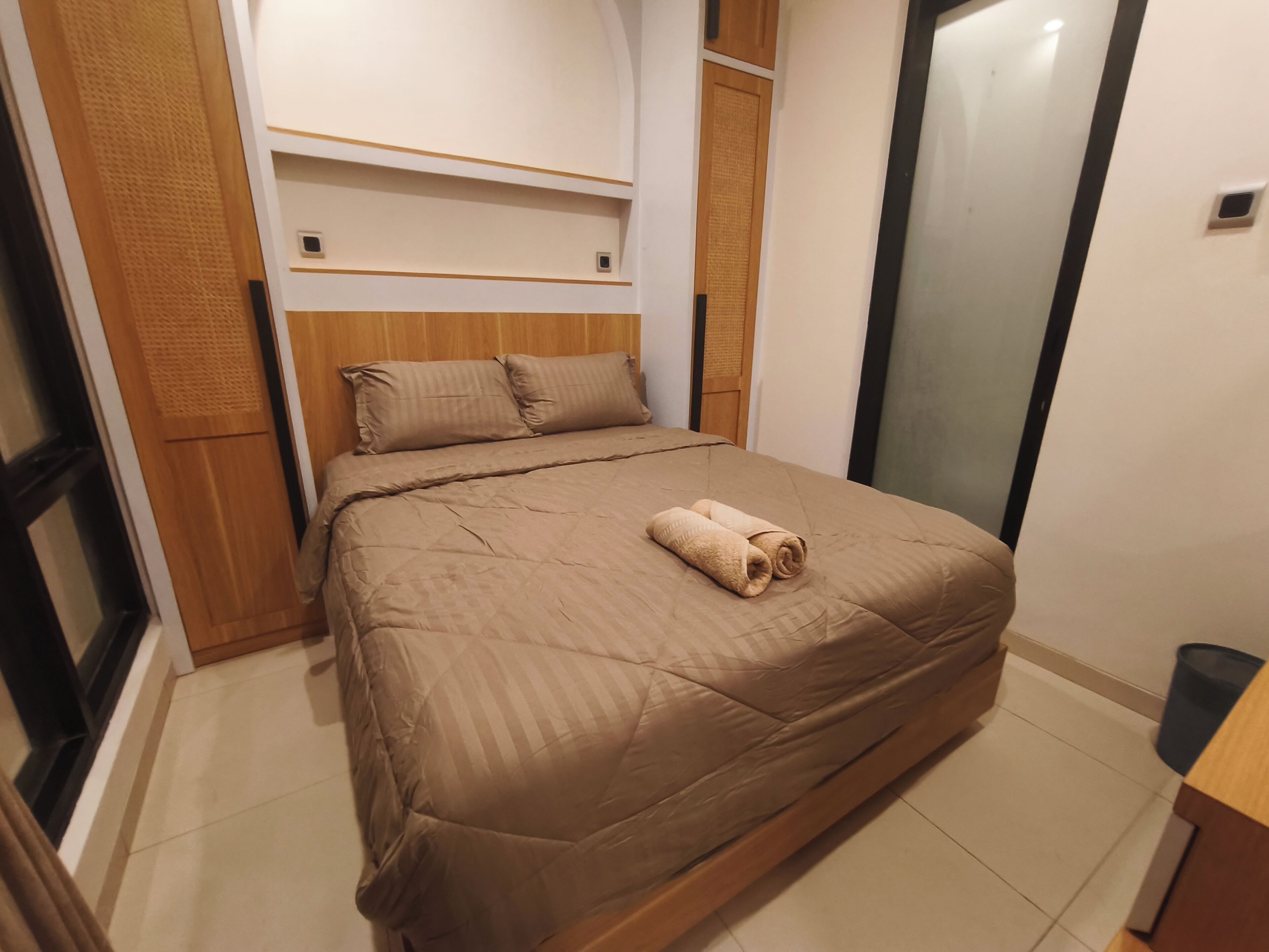 A neatly made bed with a brown bedspread and pillows, a wooden headboard, and a small wooden shelf above the bed.