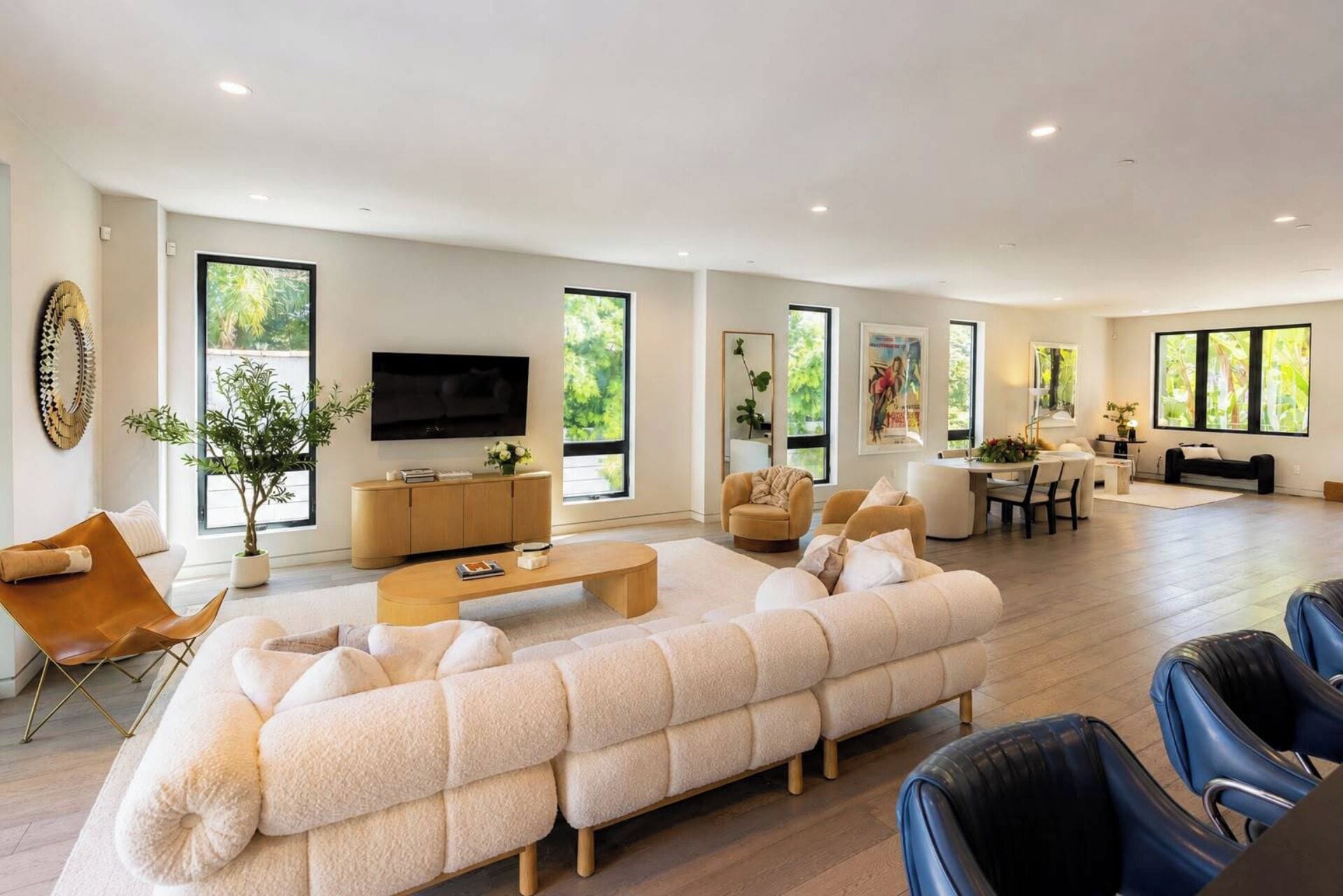 A modern living room with a large sofa, a coffee table, and a TV. There are large windows and a dining area with chairs.