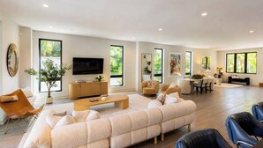 Ein modernes Wohnzimmer mit einer großen Couch, einem Couchtisch und einem Fernseher. Große Fenster und ein Essbereich mit Stühlen sind ebenfalls vorhanden.